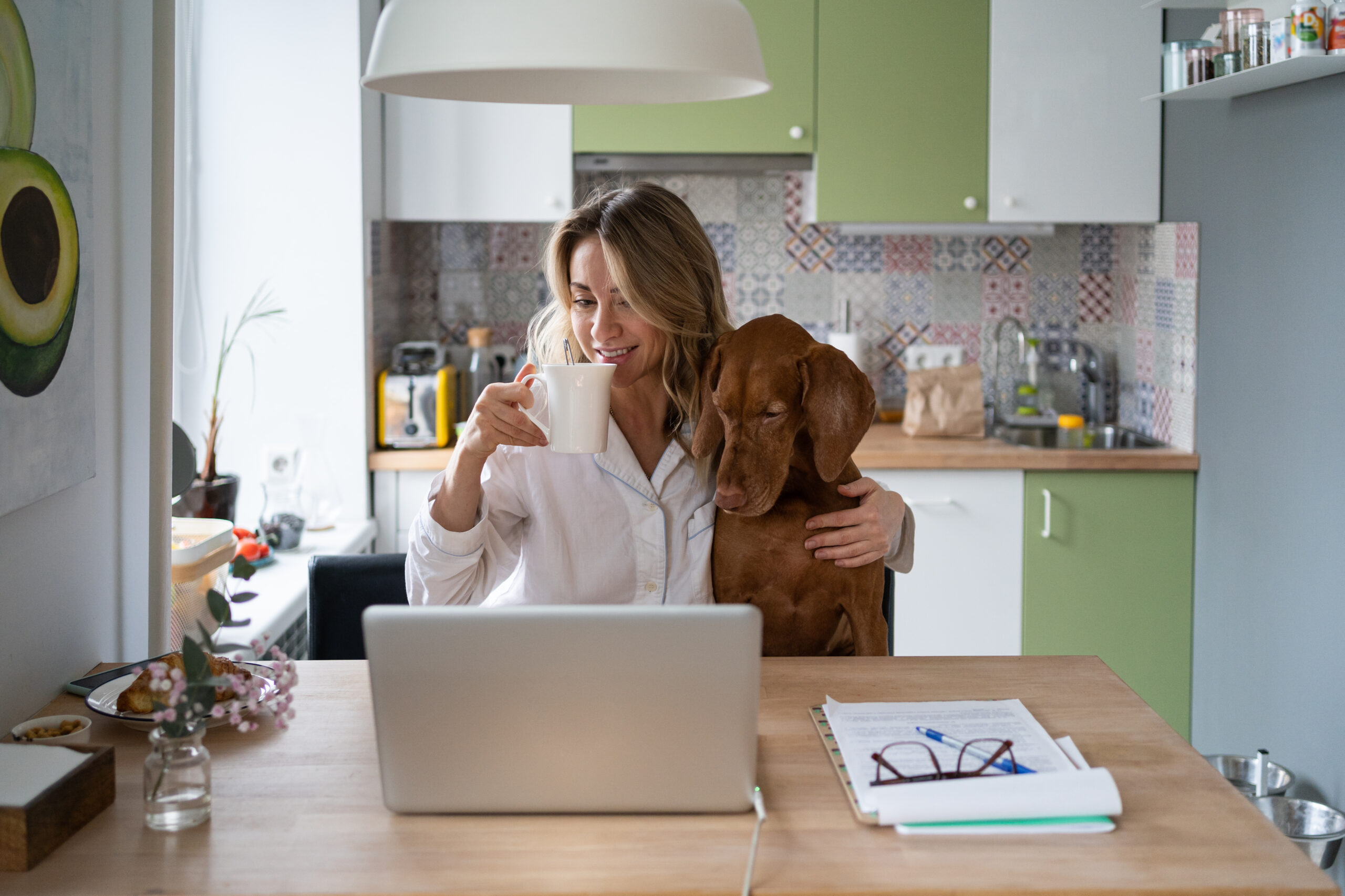 Young woman in pjs sits with her morning coffee and dog, looking at the laptop