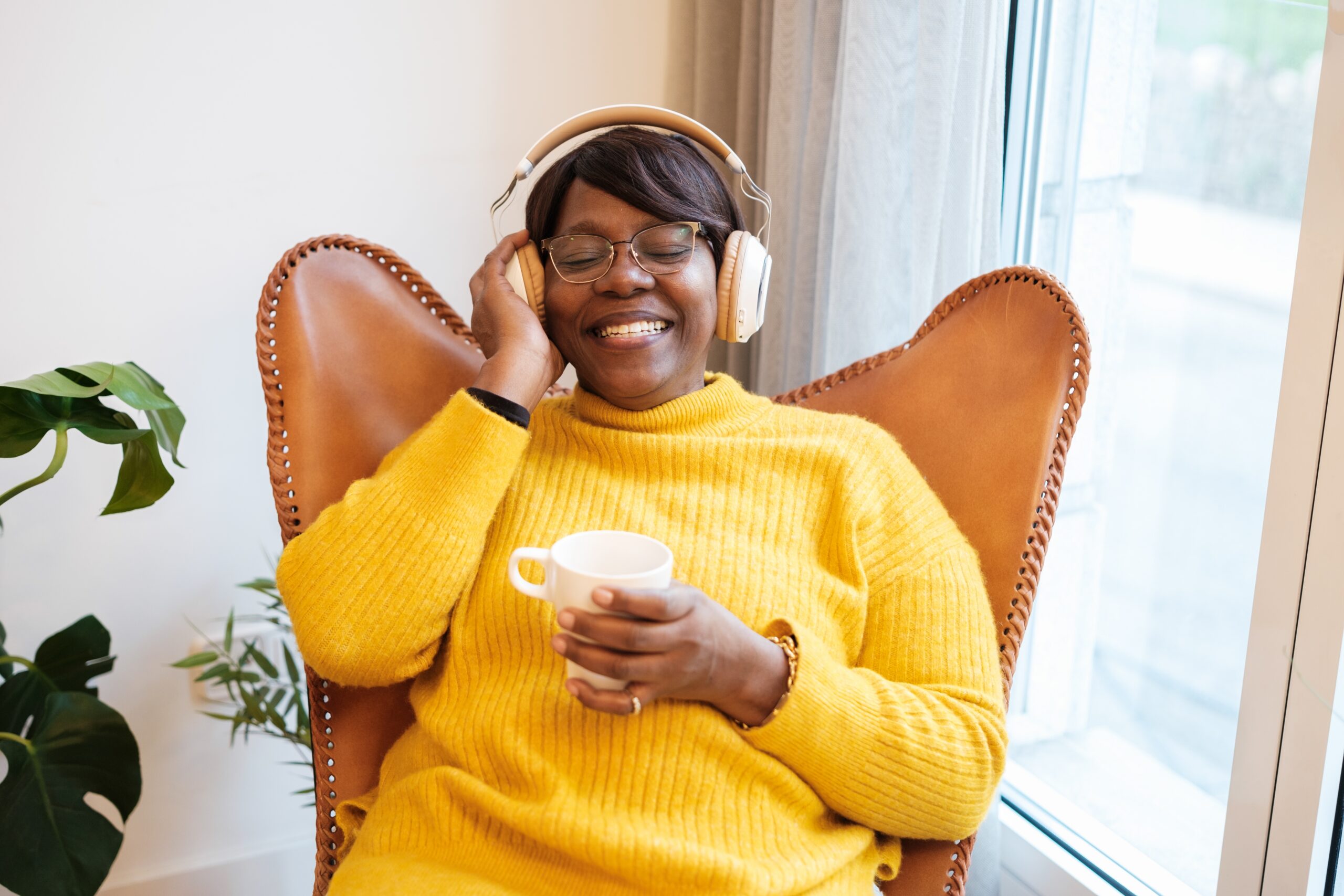 Lady sits in a comfortable chair, happily listening to music on her headphones with a cup of tea