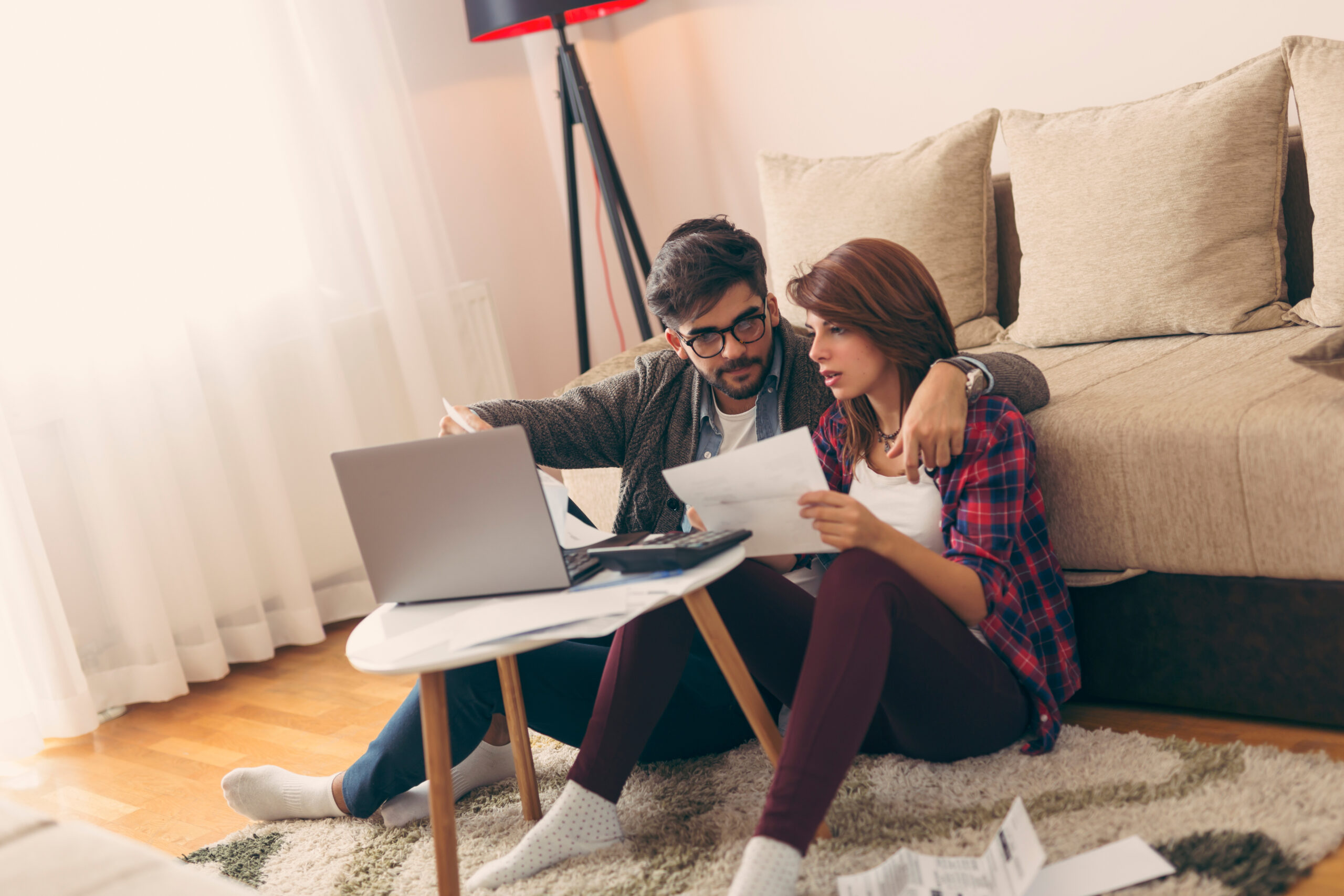 Young couple sit on floor and ponder over bills with laptop