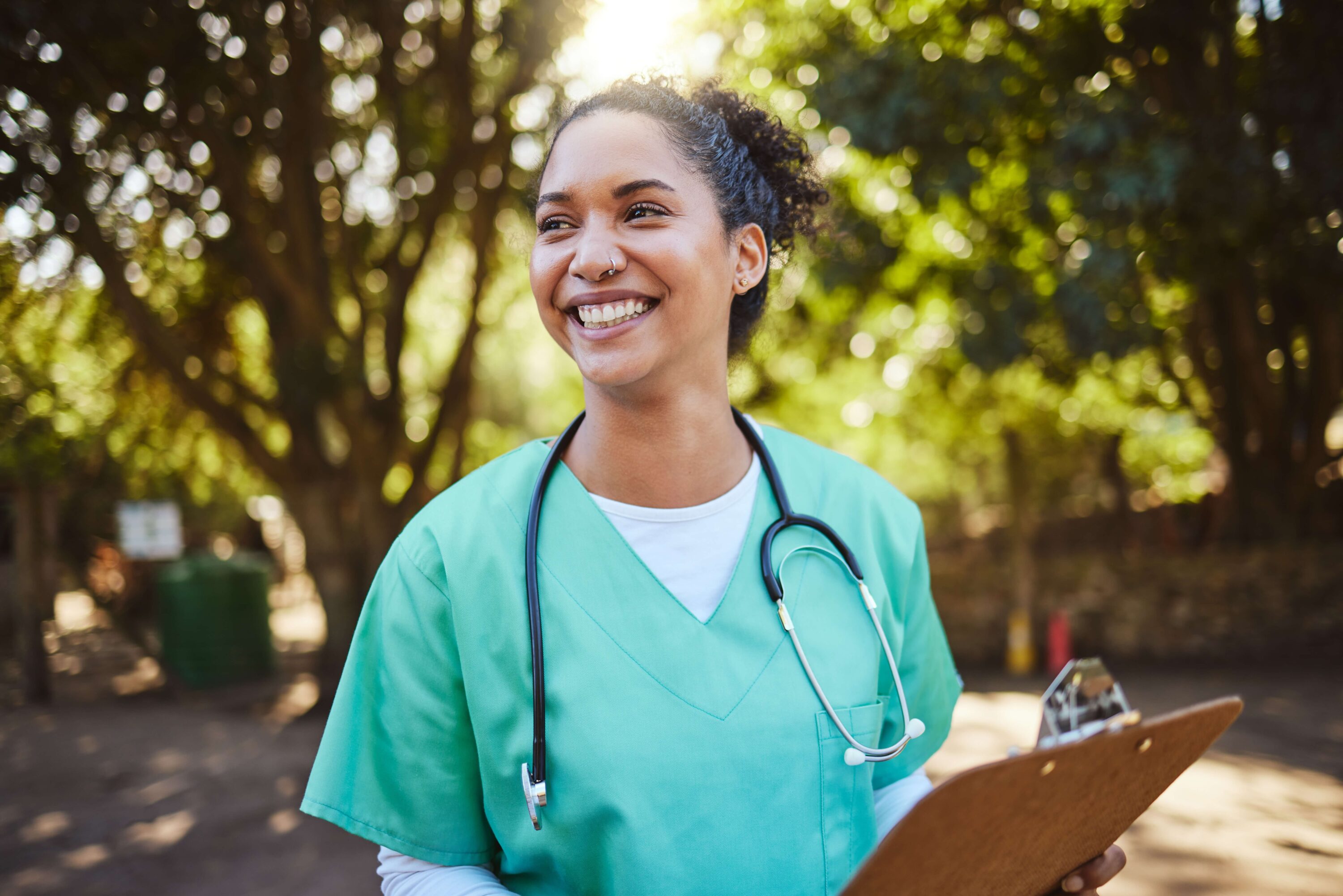 District nurse holding a clipboard and wearing pale green scrubs. She's smiling outside with trees behind her and the sun shining through them.