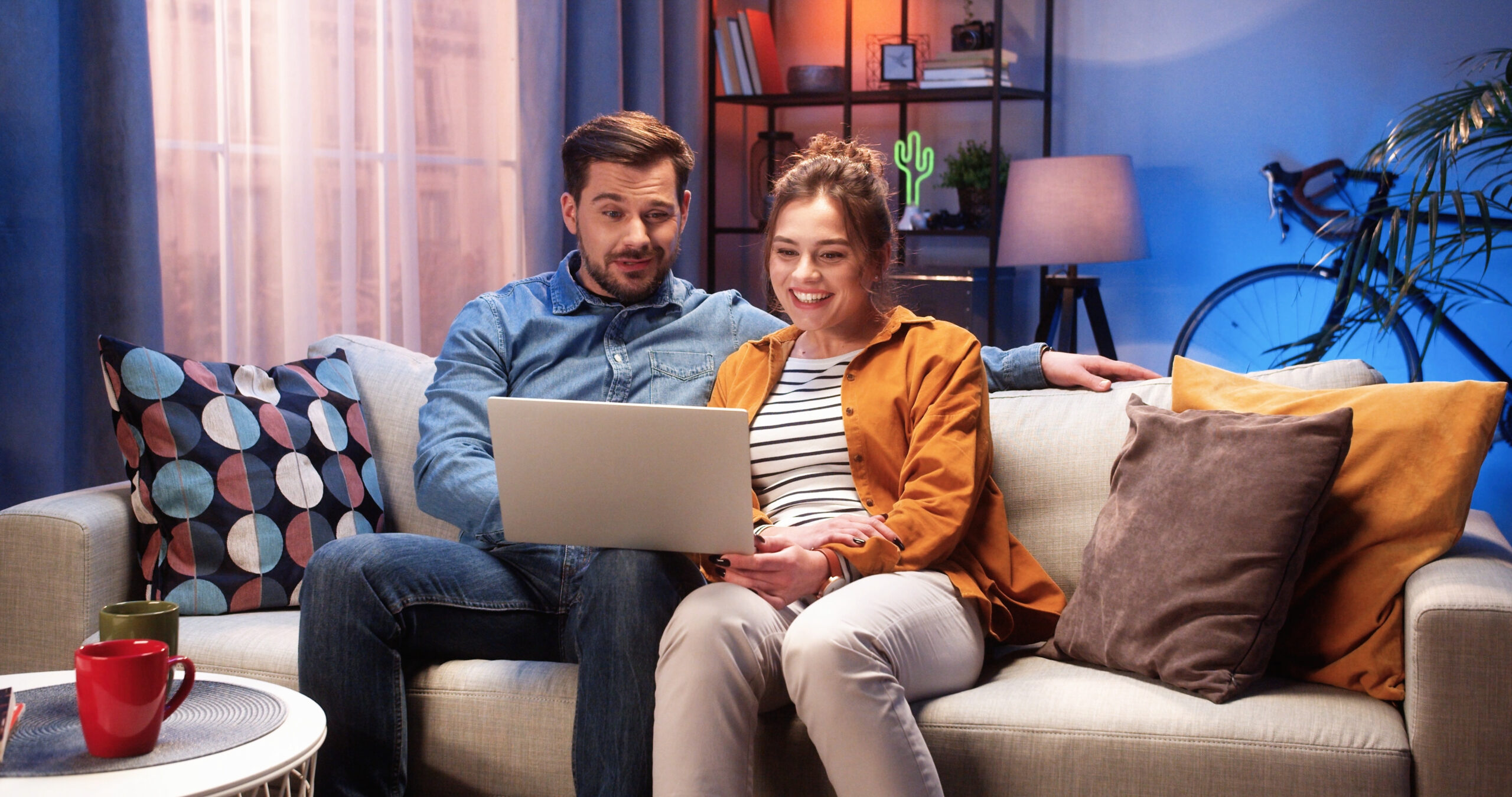 Young couple sit on sofa and smile at laptop