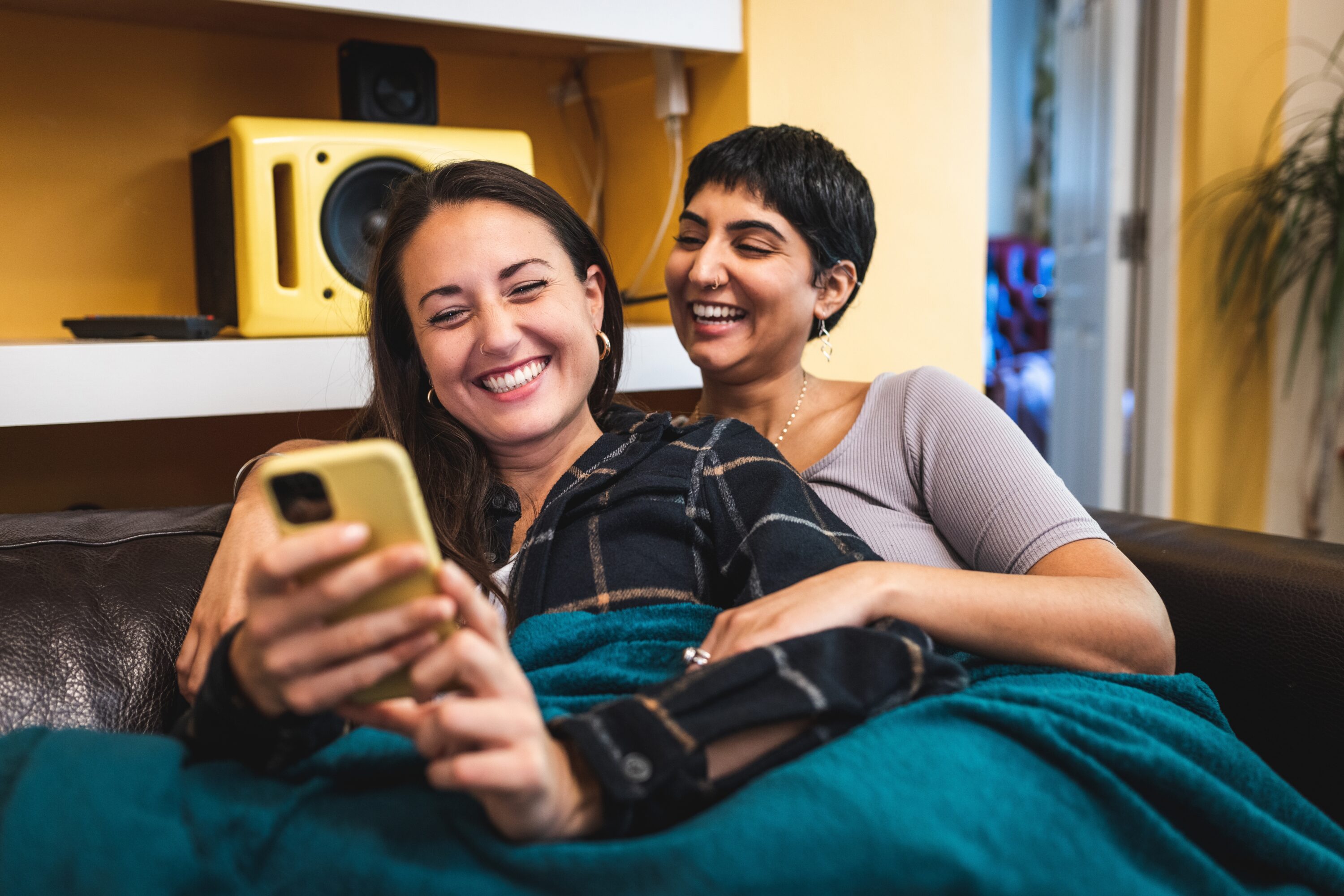 Young lesbian couple cuddling on the sofa, looking at their phones and smiling