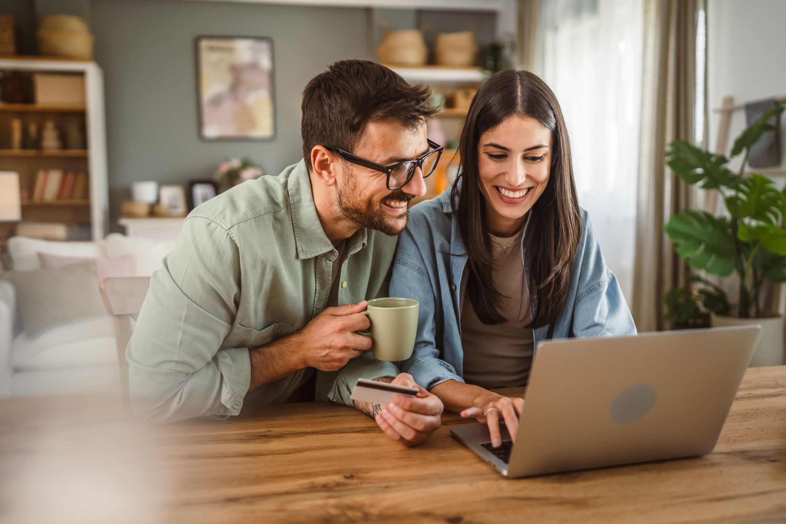 Young adult couple excitedly browsing on the laptop at home, with a coffee and a credit card in their hands