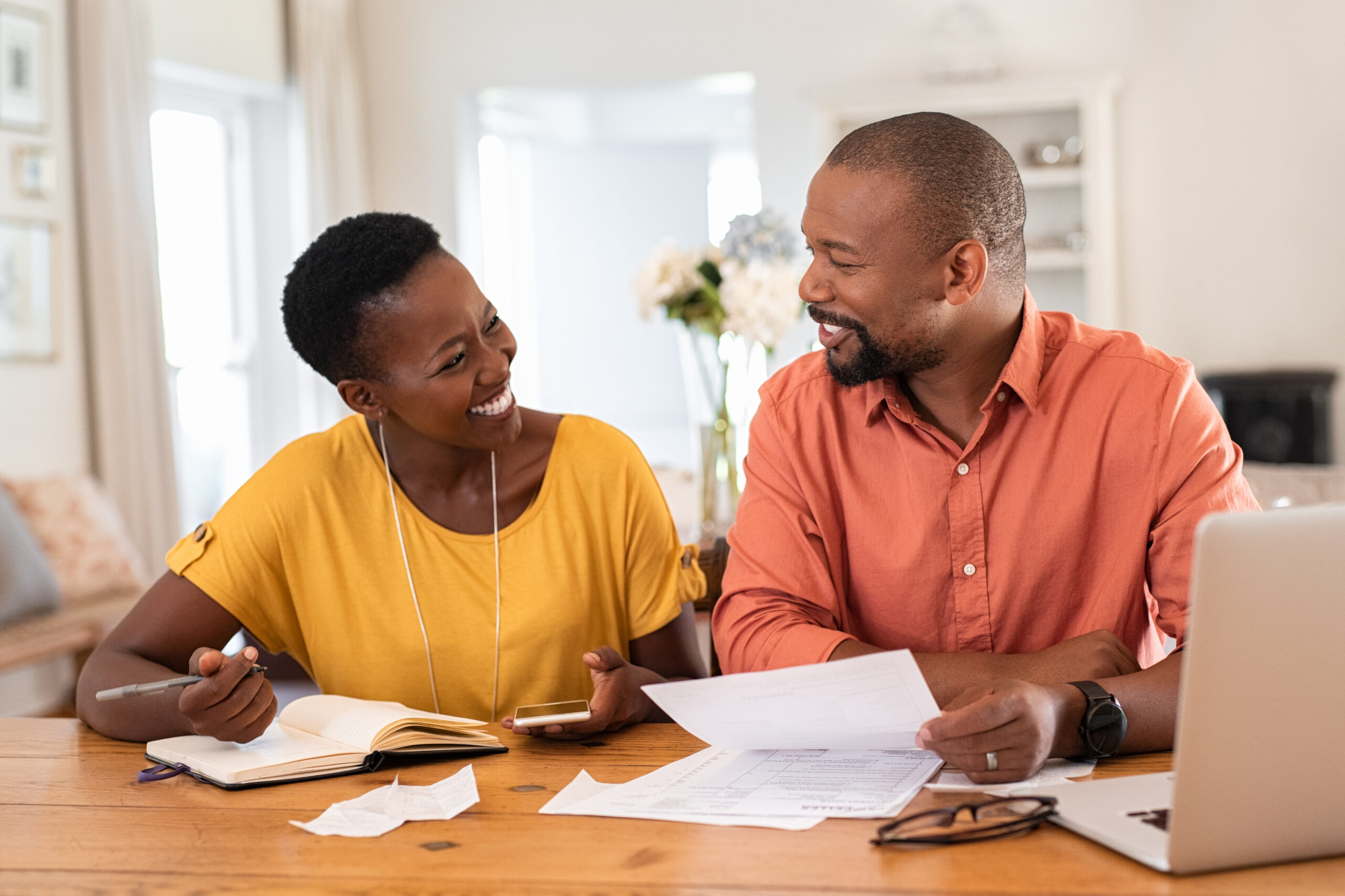 Couple sit at home with a laptop and paperwork, smiling at each other as they work