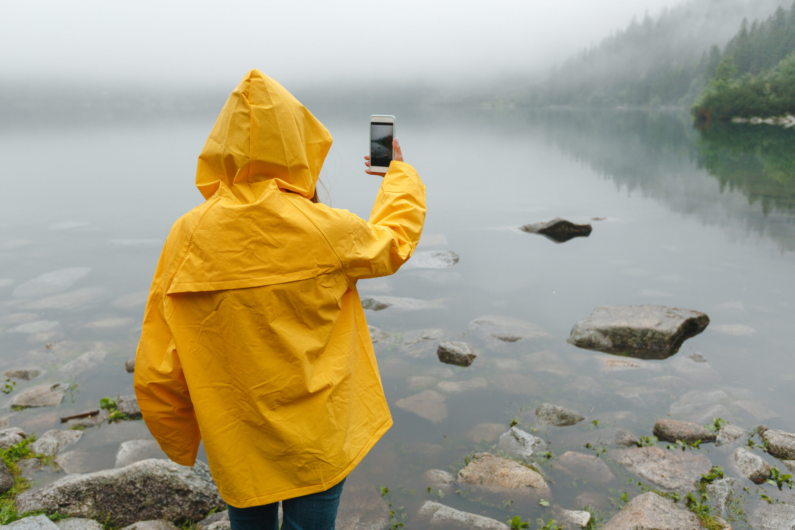 A woman in yello jacket holding her phone next to a lake