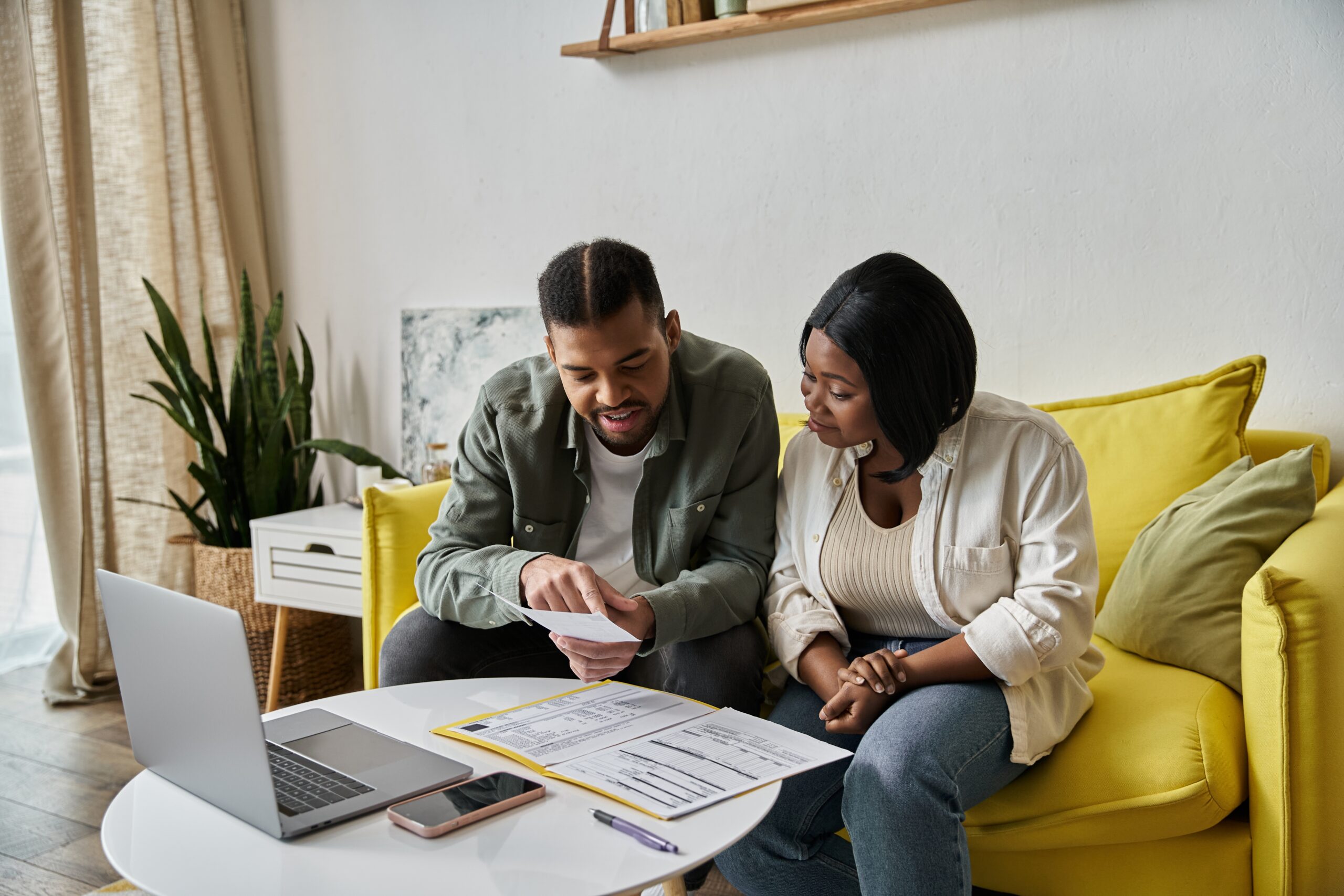 African American couple sit on a yellow sofa reviewing documents and a laptop